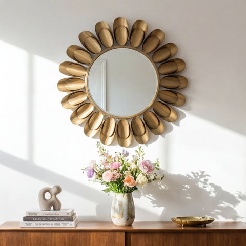Round decorative wall mirror with sculpted gold-toned petal frame above a wooden console table with flowers and minimalist decor in soft natural light.