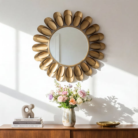 Round decorative wall mirror with sculpted gold-toned petal frame above a wooden console table with flowers and minimalist decor in soft natural light.