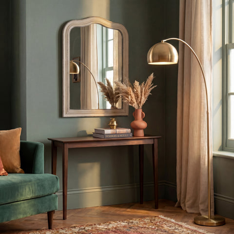 Decorative grey wall mirror with a softly curved silhouette and distressed finish, styled above a dark wood console table in a warm, elegant interior.