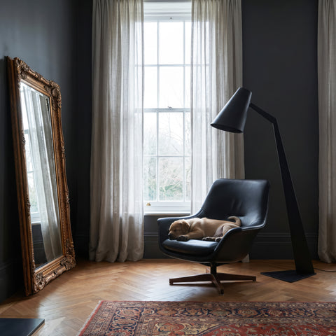 Large ornate gold full-length floor mirror leaning beside a modern black lounge chair and floor lamp in a sophisticated dark interior.