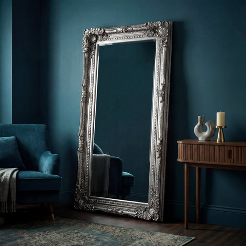 Large ornate full-length floor mirror with an antique silver carved frame leaning against a dark teal wall in an elegant modern living room.