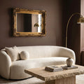Ornate rectangular wall mirror with an antique gold carved frame displayed above a curved cream sofa in a warm, elegant living room.