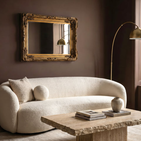 Ornate rectangular wall mirror with an antique gold carved frame displayed above a curved cream sofa in a warm, elegant living room.