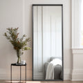 Full-length mirror with black frame leaning against a wall next to a small table with a vase of flowers.