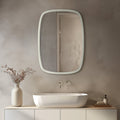 Irregular rounded rectangular wall mirror with a slim white frame displayed above a modern vessel sink in a minimalist neutral bathroom.