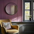 Black framed round mirror above a yellow armchair with a pillow in a room with purple paneled walls and a window.