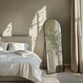 Bedroom with a large cheval black framed mirror reflecting a plant, draped curtains, and a neutral colour palette.
