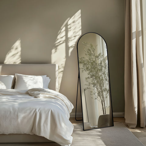 Bedroom with a large cheval black framed mirror reflecting a plant, draped curtains, and a neutral colour palette.