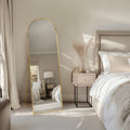 Neatly arranged bedroom with a free standing gold mirror, bed, and side table.