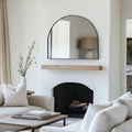 Arched black wall mirror with a slim frame, styled above a fireplace mantel to add contrast and understated elegance to a calm neutral interior.