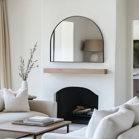 Arched black wall mirror with a slim frame, styled above a fireplace mantel to add contrast and understated elegance to a calm neutral interior.