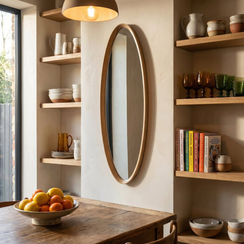 Oval wall mirror with a natural wood frame displayed in a warm modern kitchen dining space with open wooden shelving and soft ambient lighting.