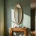 Oval wall mirror with a natural wood frame mounted above a ribbed wooden console table in a warm minimalist interior with soft sunlight.