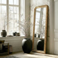Ornate gold leaner mirror with an intricately carved frame, styled beside oversized vases and soft natural light for an elegant, timeless room setting.