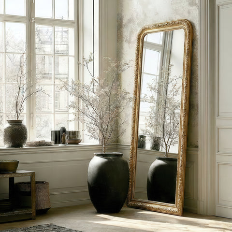 Ornate gold leaner mirror with an intricately carved frame, styled beside oversized vases and soft natural light for an elegant, timeless room setting.